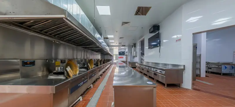 A long stainless-steel exhaust hood running above a line of cooking stations in a large commercial kitchen