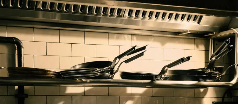 A restaurant kitchen exhaust hood with grease filters above a tiled wall and a shelf of frying pans
