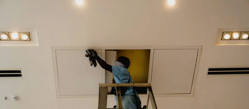 A technician on a ladder reaching through an open ceiling access panel